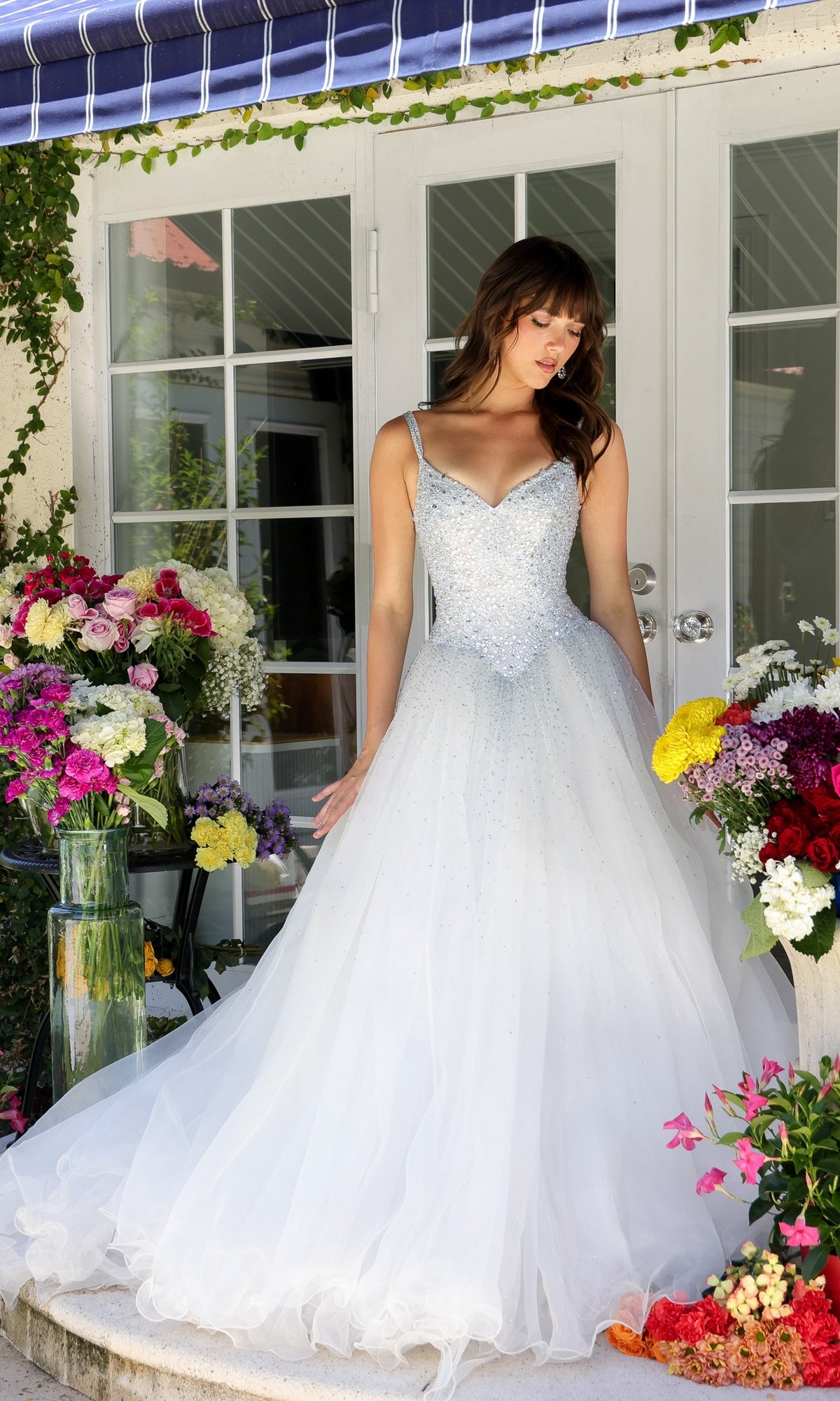 A woman wears the Ava Presley Long Prom Dress 29522—a white, sparkling ball gown with train—as she stands before glass doors, surrounded by vibrant floral arrangements on either side.