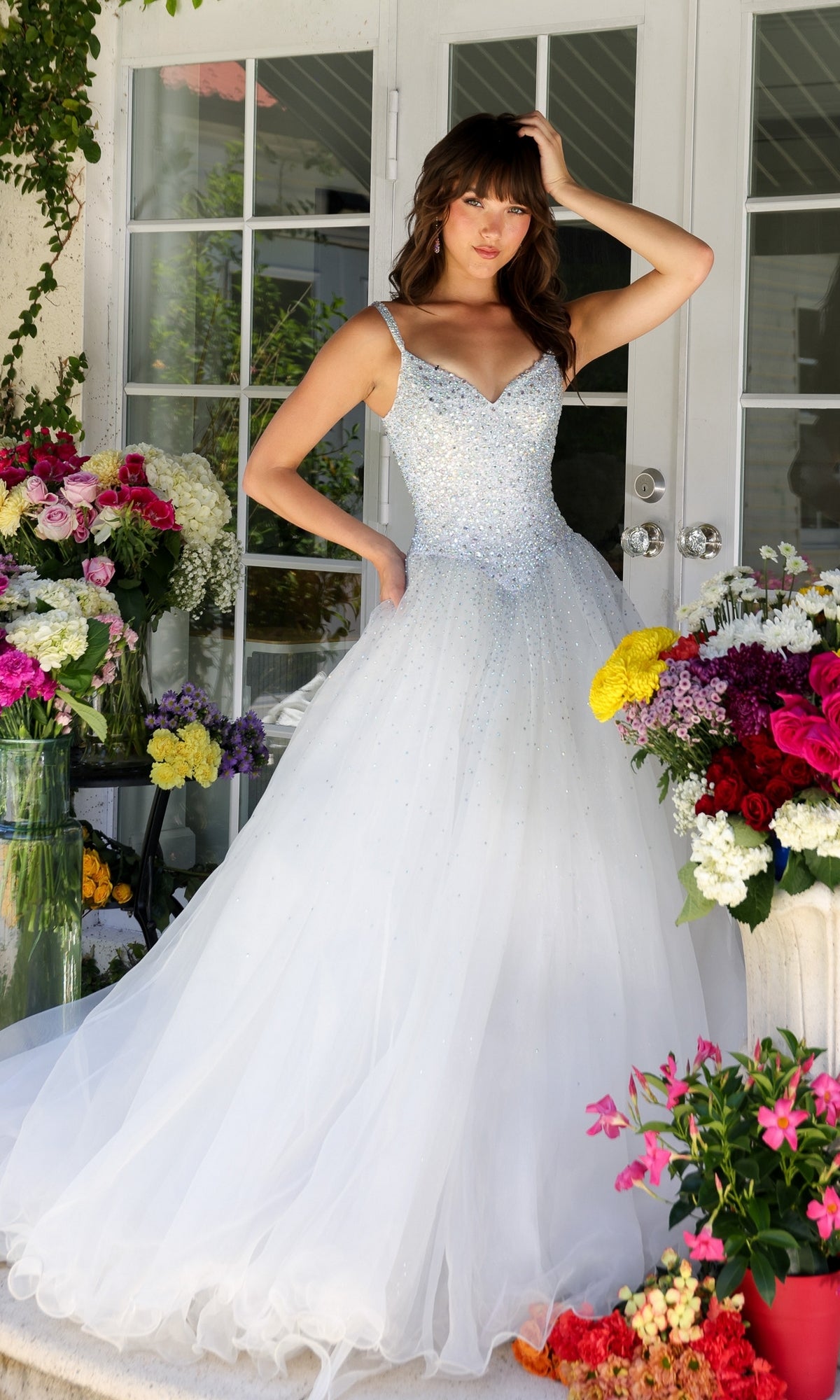 A woman wearing the Long Prom Dress 29522 by Ava Presley, a sparkly white ball gown with a sweetheart neckline, stands among colorful floral arrangements in front of glass doors.