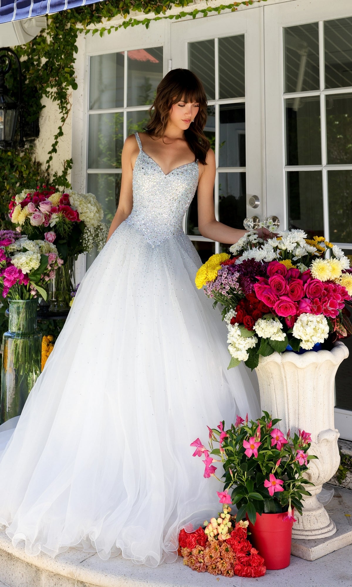 A woman in the Long Prom Dress 29522 by Ava Presley, a white sparkling A-line ball gown with a sweetheart neckline, stands on a porch next to large bouquets of colorful flowers in vases and pots.