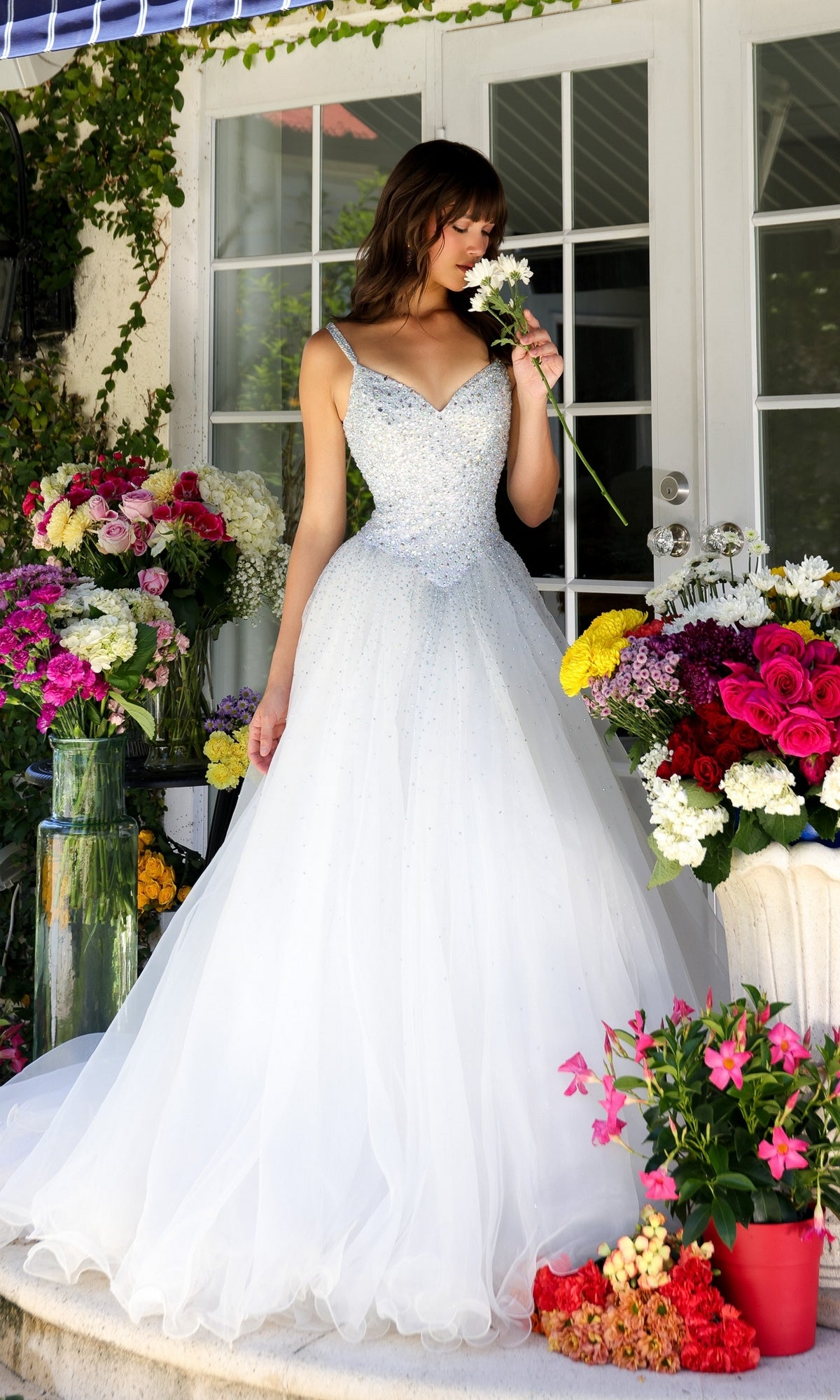 A woman wearing the Long Prom Dress 29522 by Ava Presley, a sparkling white ball gown with a train, stands among vibrant flowers and smells a white blossom in front of glass doors.