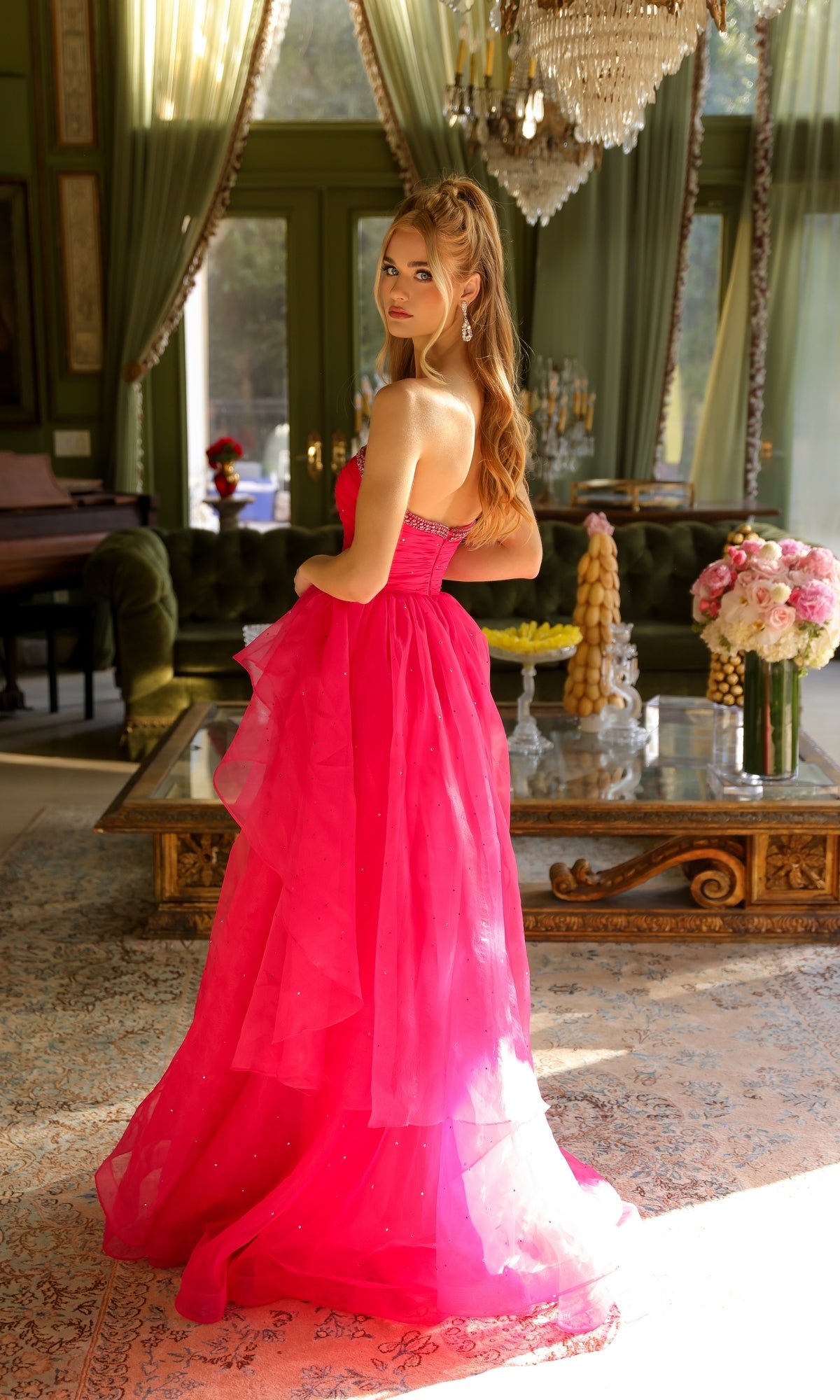A woman in the Ava Presley 29524 strapless long prom ball gown poses indoors, glancing over her shoulder in a room with green walls, large windows, ornate curtains, and a chandelier.