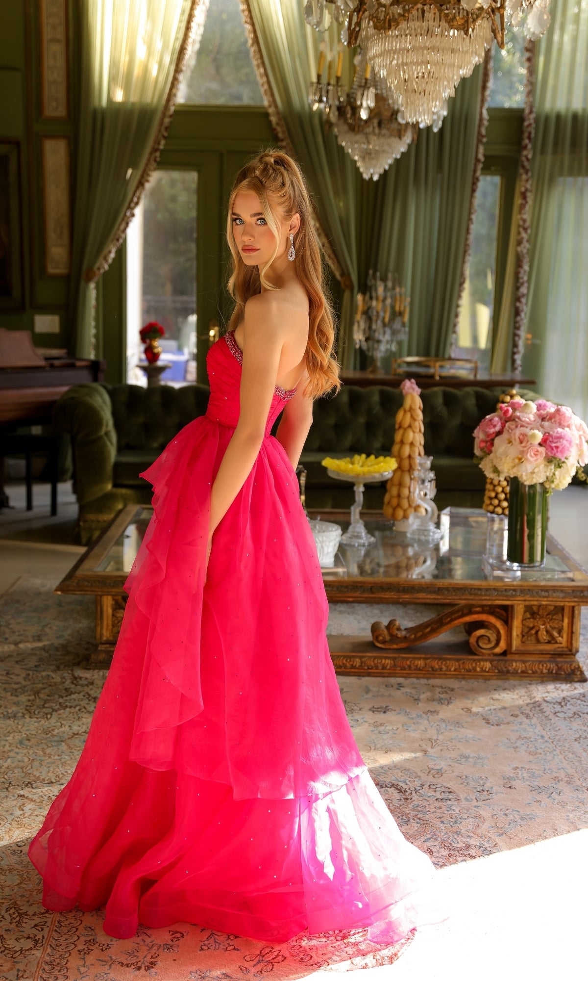 Wearing the Strapless Long Prom Ball Gown: Ava Presley 29524 in bright pink, a woman stands in a luxurious living room with green walls, a chandelier, and elegant floral arrangements.