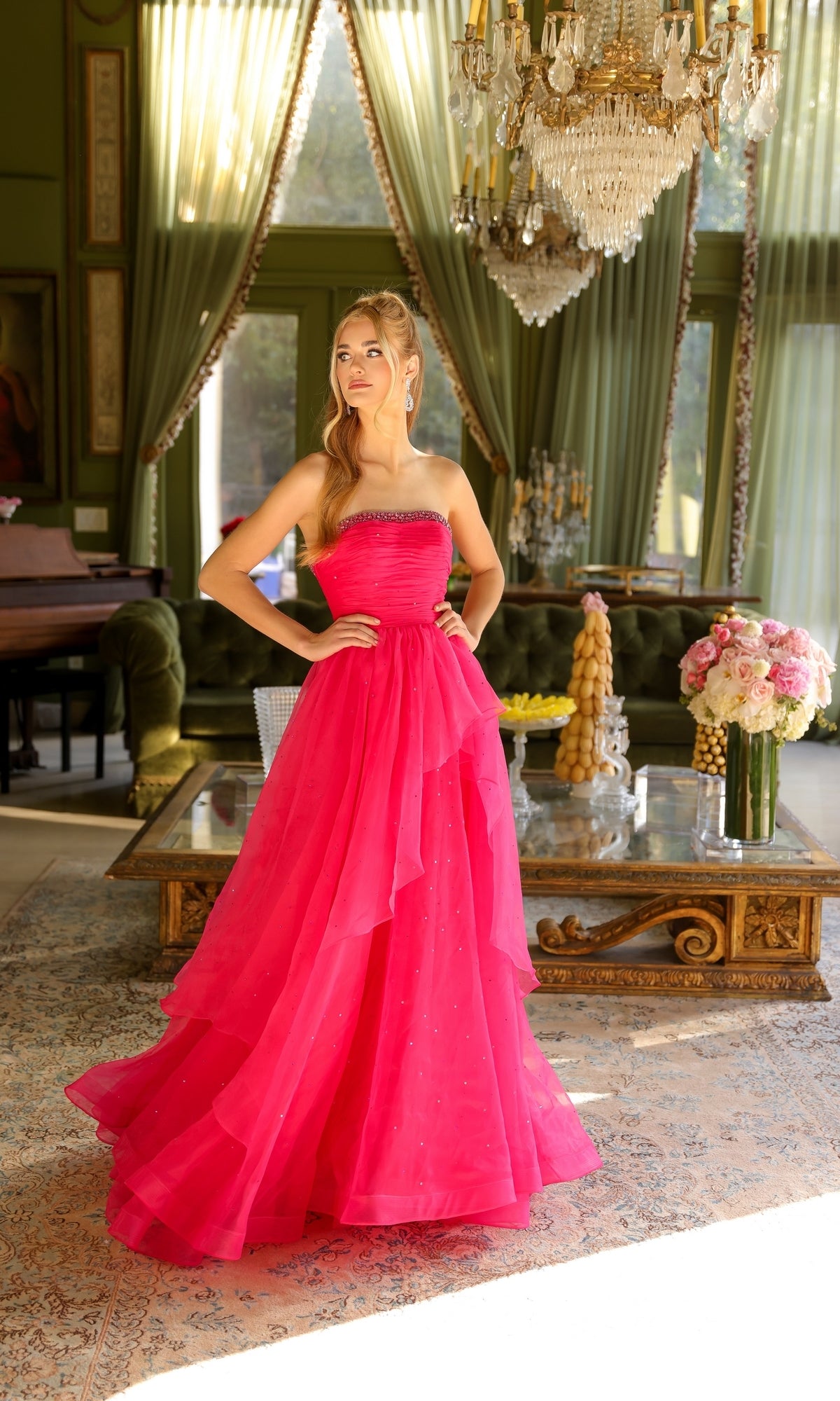 A woman wearing the Ava Presley 29524 strapless long prom ball gown in bright pink stands in an elegant, sunlit room with large windows, green furniture, flowers, and a chandelier.