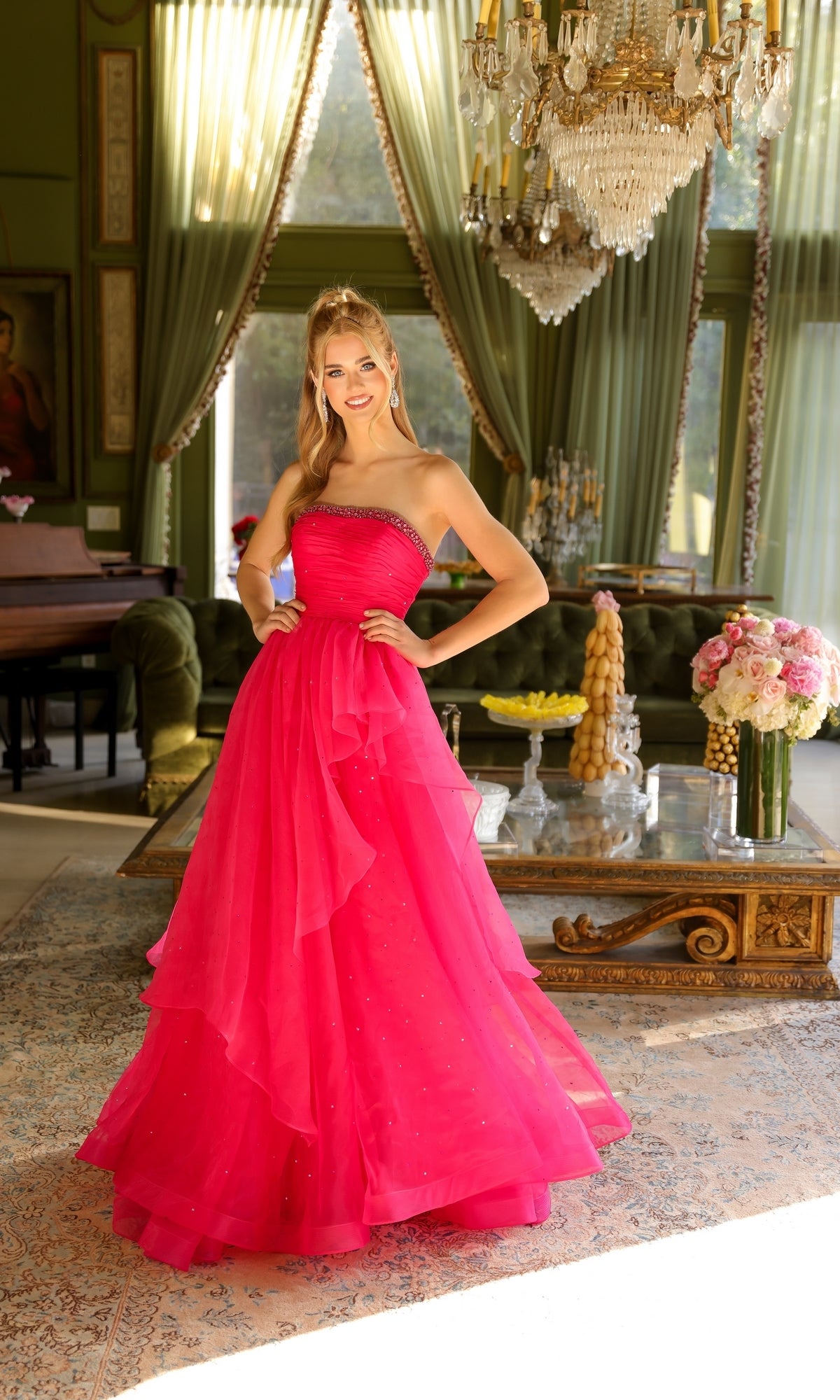A woman stands in an elegant room, smiling at the camera with her hands on her hips, wearing the Ava Presley 29524 Strapless Long Prom Ball Gown in bright pink.