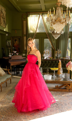 A woman wears the Ava Presley 29524 Strapless Long Prom Ball Gown in bright pink, standing in an elegant, sunlit room with chandeliers, vintage furniture, and large windows.