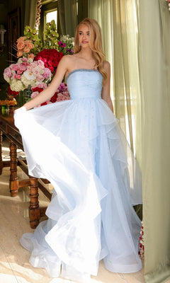 A woman in the Ava Presley 29524 Strapless Long Prom Ball Gown, light blue and a-line style, stands indoors by a window with green curtains, holding her dress beside a table topped with a large bouquet of flowers.