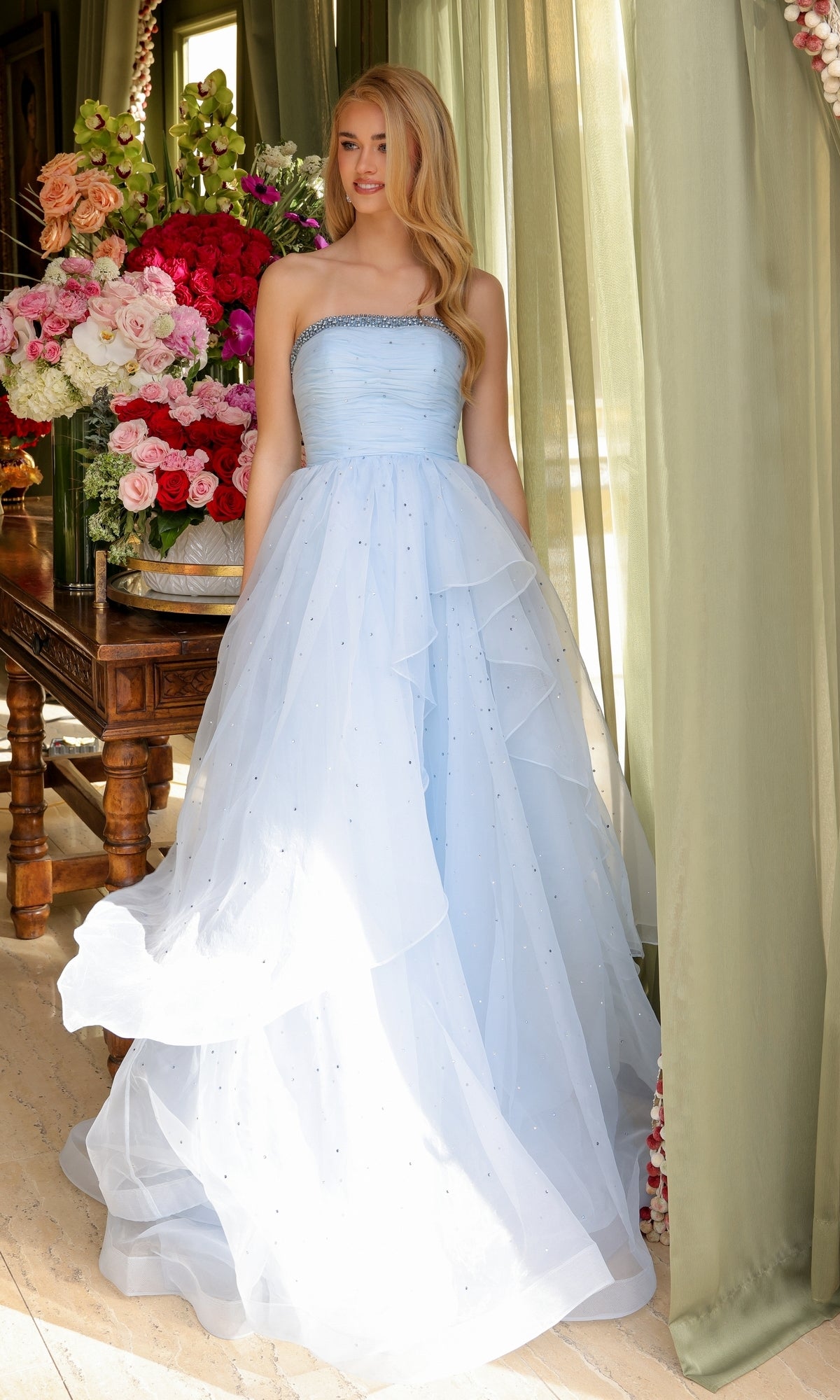 A woman in the Ava Presley 29524 Strapless Long Prom Ball Gown, light blue with layered tulle, stands by a window framed with green curtains and floral arrangements.
