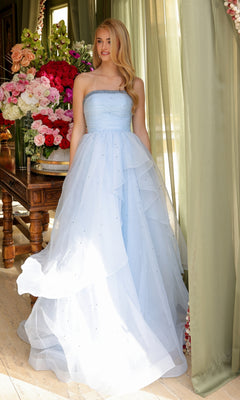 A woman in the Ava Presley 29524 Strapless Long Prom Ball Gown, light blue with layered tulle, stands by a window framed with green curtains and floral arrangements.