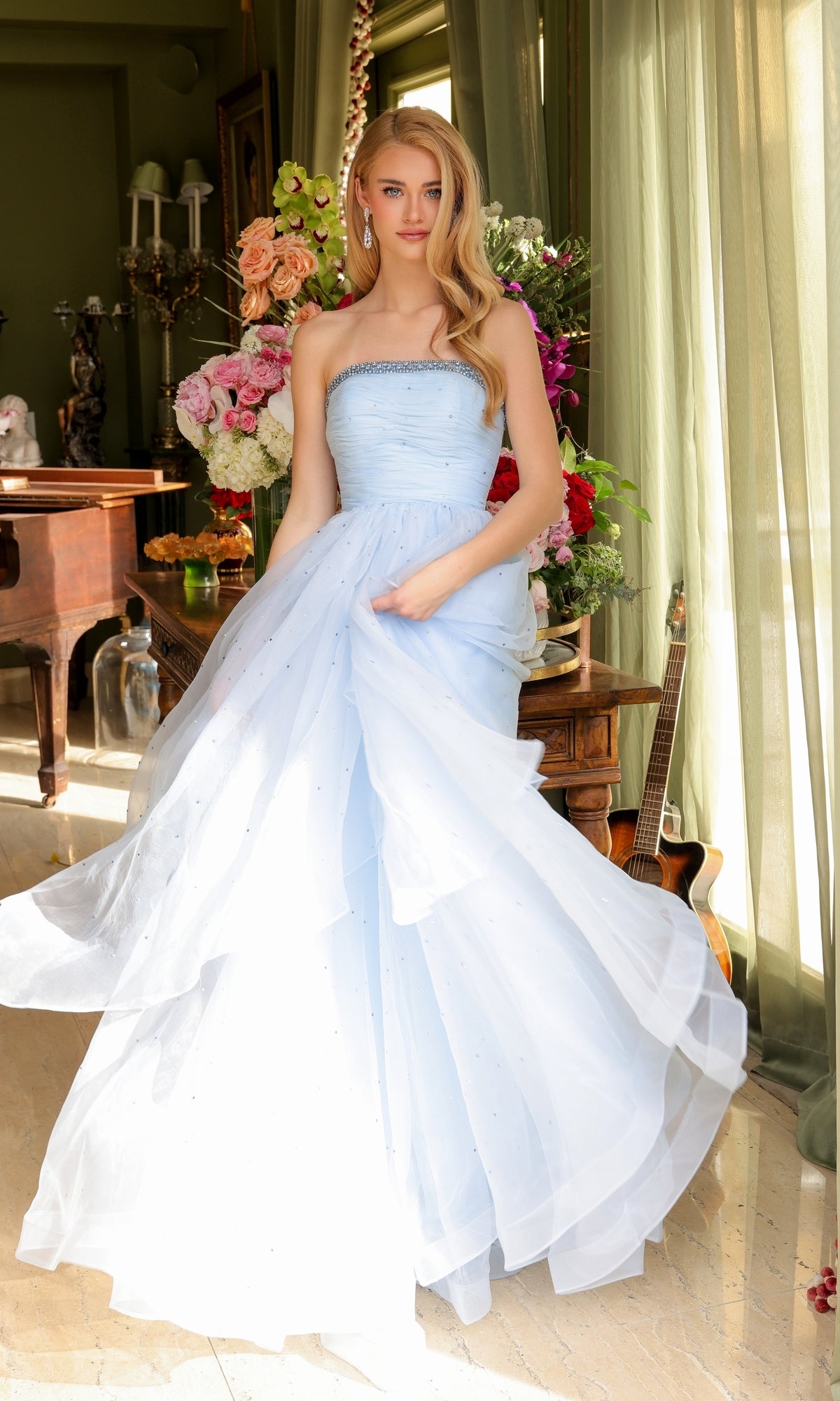 A woman wearing the Ava Presley 29524 Strapless Long Prom Ball Gown stands indoors by a large flower bouquet and a guitar, sunlight streaming through green curtains.