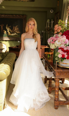 A woman in the Ava Presley 29524 Strapless Long Prom Ball Gown stands indoors by a green couch and a table with a large bouquet of pink and red flowers.