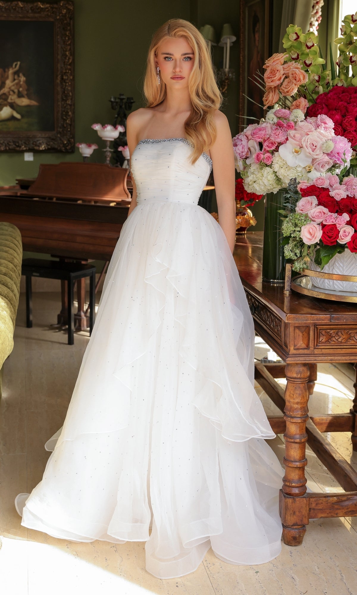 A woman wearing the Ava Presley 29524 Strapless Long Prom Ball Gown stands indoors by a wooden table with large floral arrangements and a piano in the background.