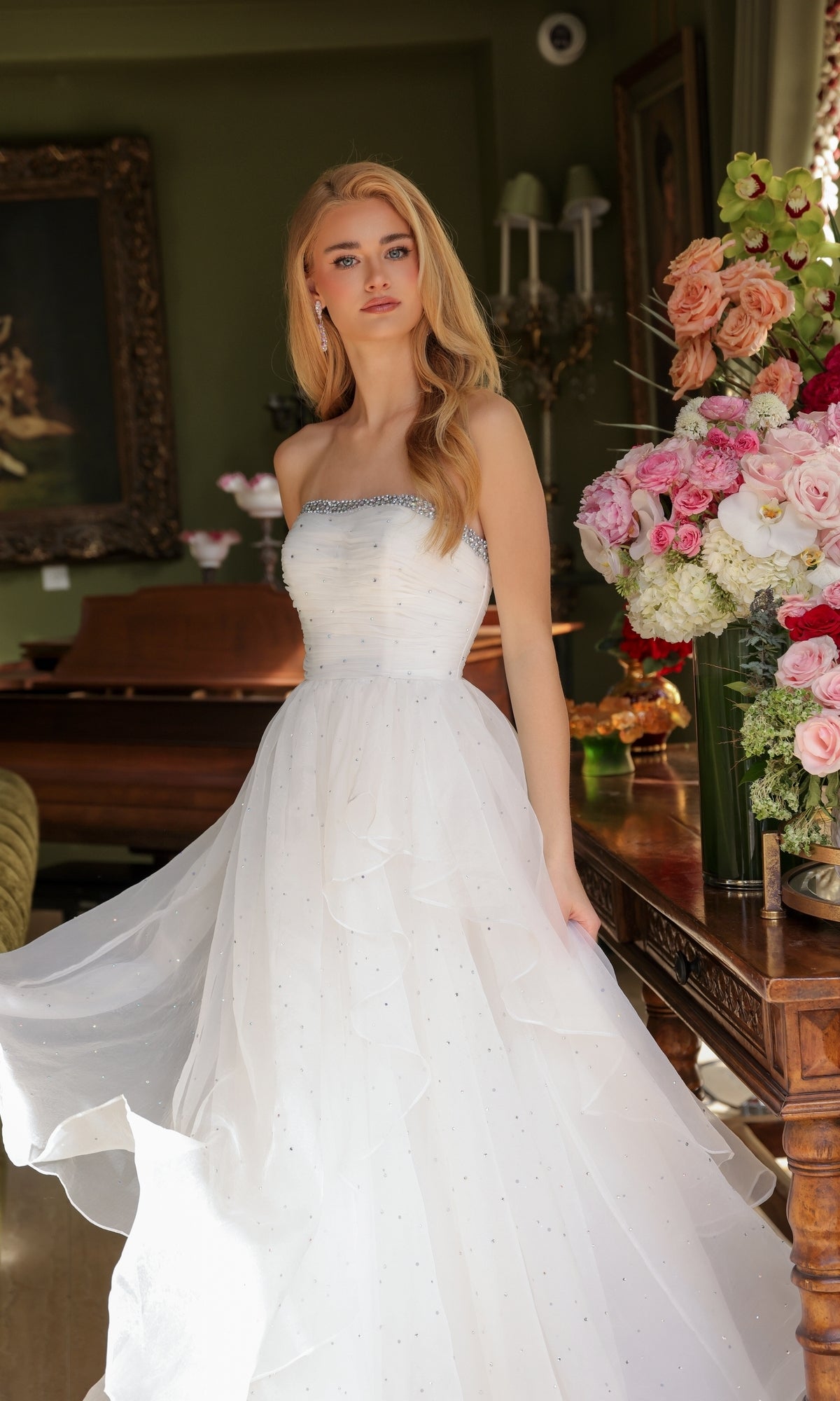 A woman in the Ava Presley 29524 strapless long prom ball gown with a beaded neckline stands indoors beside a wooden table topped with a large floral arrangement.