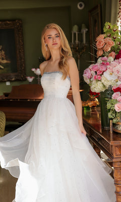 A woman in the Ava Presley 29524 strapless long prom ball gown with a beaded neckline stands indoors beside a wooden table topped with a large floral arrangement.