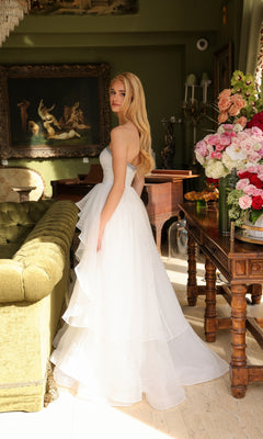 A woman in the Ava Presley 29524 strapless long prom ball gown stands beside a wooden table with a large flower arrangement in an elegantly decorated room.