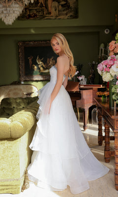 Wearing the Ava Presley 29524 Strapless Long Prom Ball Gown, a woman stands beside a green sofa and wooden piano in a vintage-style room with floral arrangements and framed artwork.