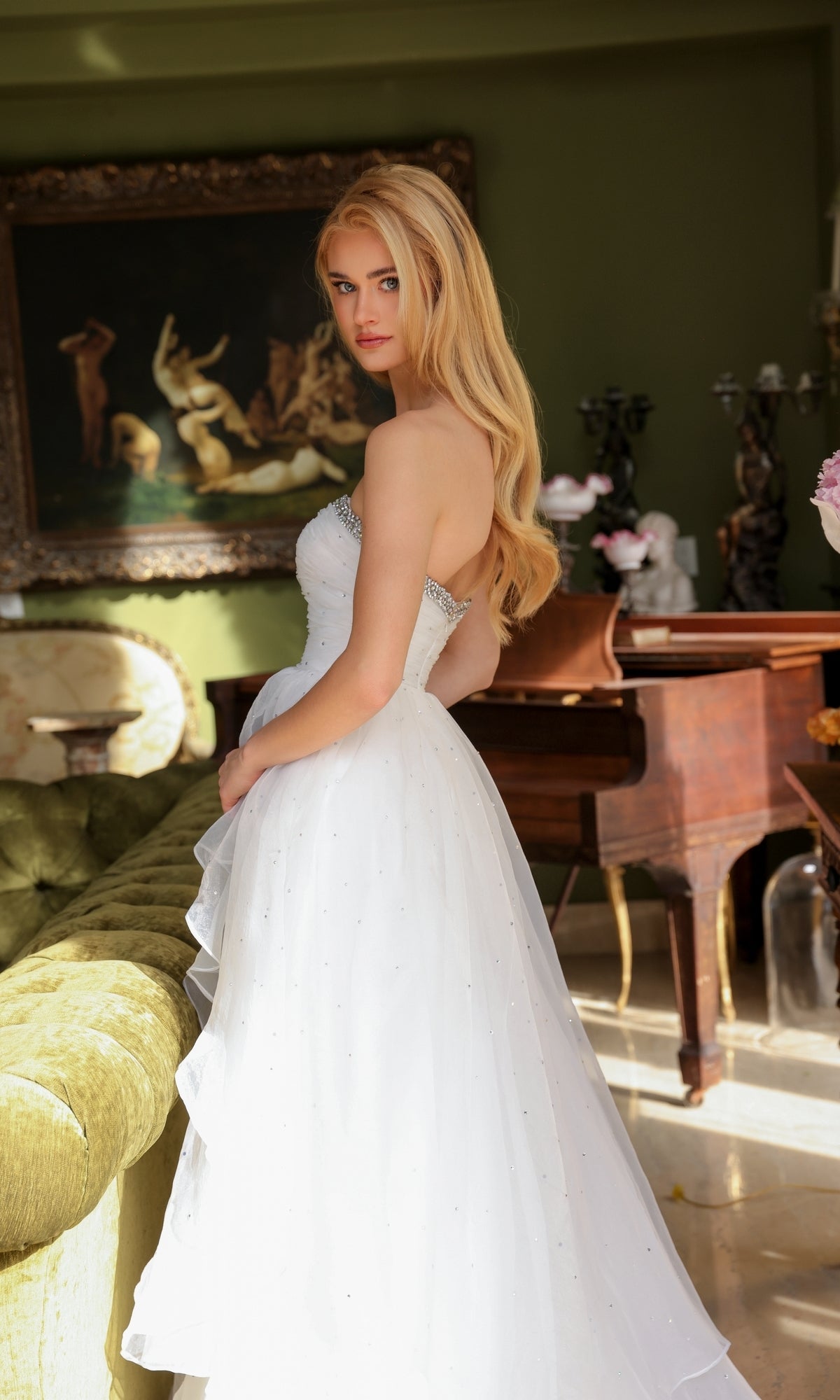 A woman with long blonde hair wears the Ava Presley 29524 Strapless Long Prom Ball Gown, standing indoors by a green sofa and piano.