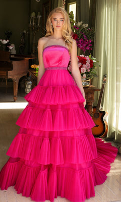 A woman stands indoors wearing the Long Prom Dress 29529 by Ava Presley, a strapless bright pink A-line gown with a beaded waist and layered ruffle ball gown skirt. Behind her are a piano, floral arrangements, and an acoustic guitar.