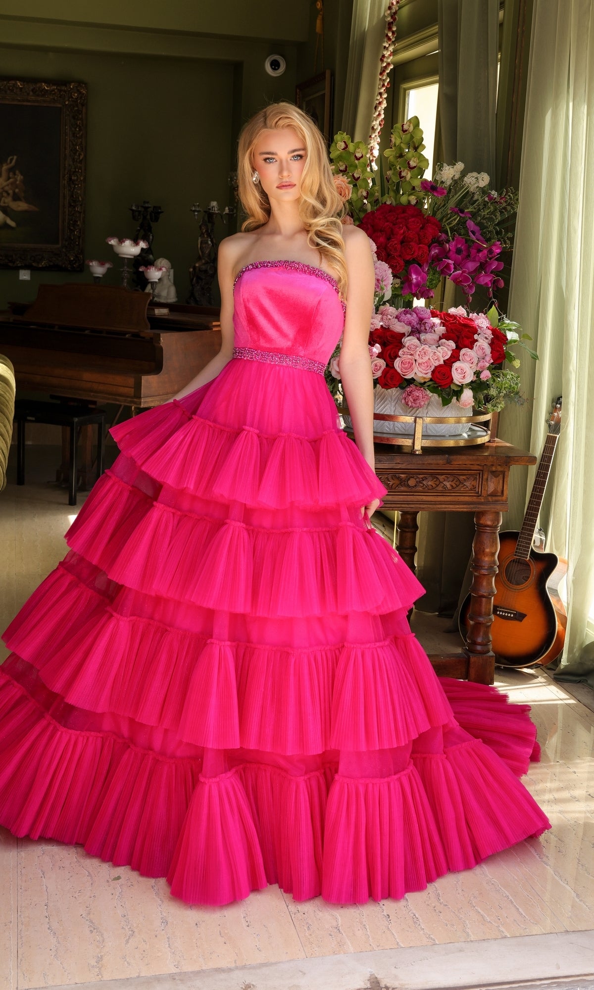 A woman indoors wears the Long Prom Dress 29529 by Ava Presley, a strapless bright pink layered ruffle ball gown, standing beside a flower-adorned table with a guitar resting against the wall.