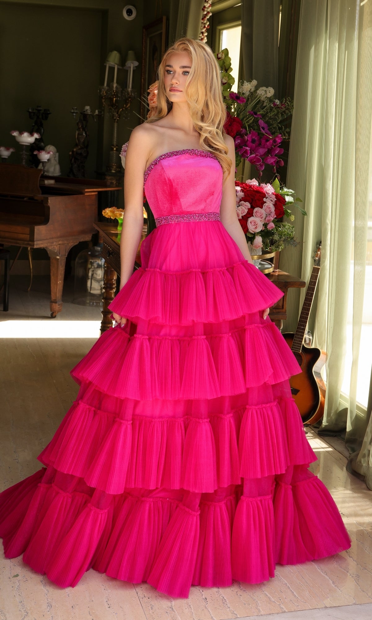 A person stands indoors wearing the Long Prom Dress 29529 by Ava Presley, a strapless bright pink A-line dress with pleated layers, surrounded by flowers and musical instruments.