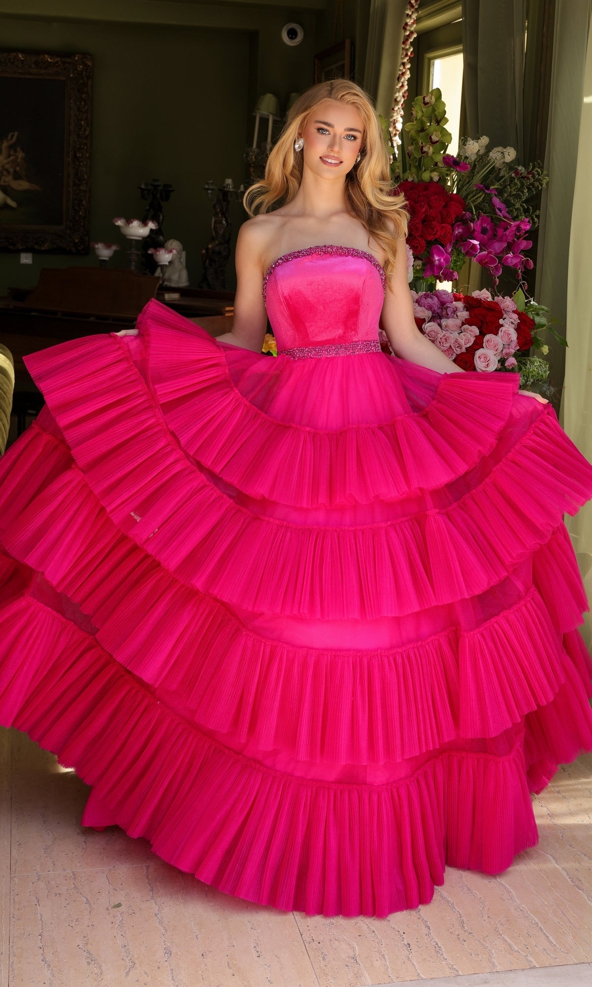 Indoors, someone showcases the Long Prom Dress 29529 by Ava Presley—a bright pink, strapless a-line gown featuring a beaded waist and layered ruffle ball gown skirt. Flowers and decorative items appear in the background.