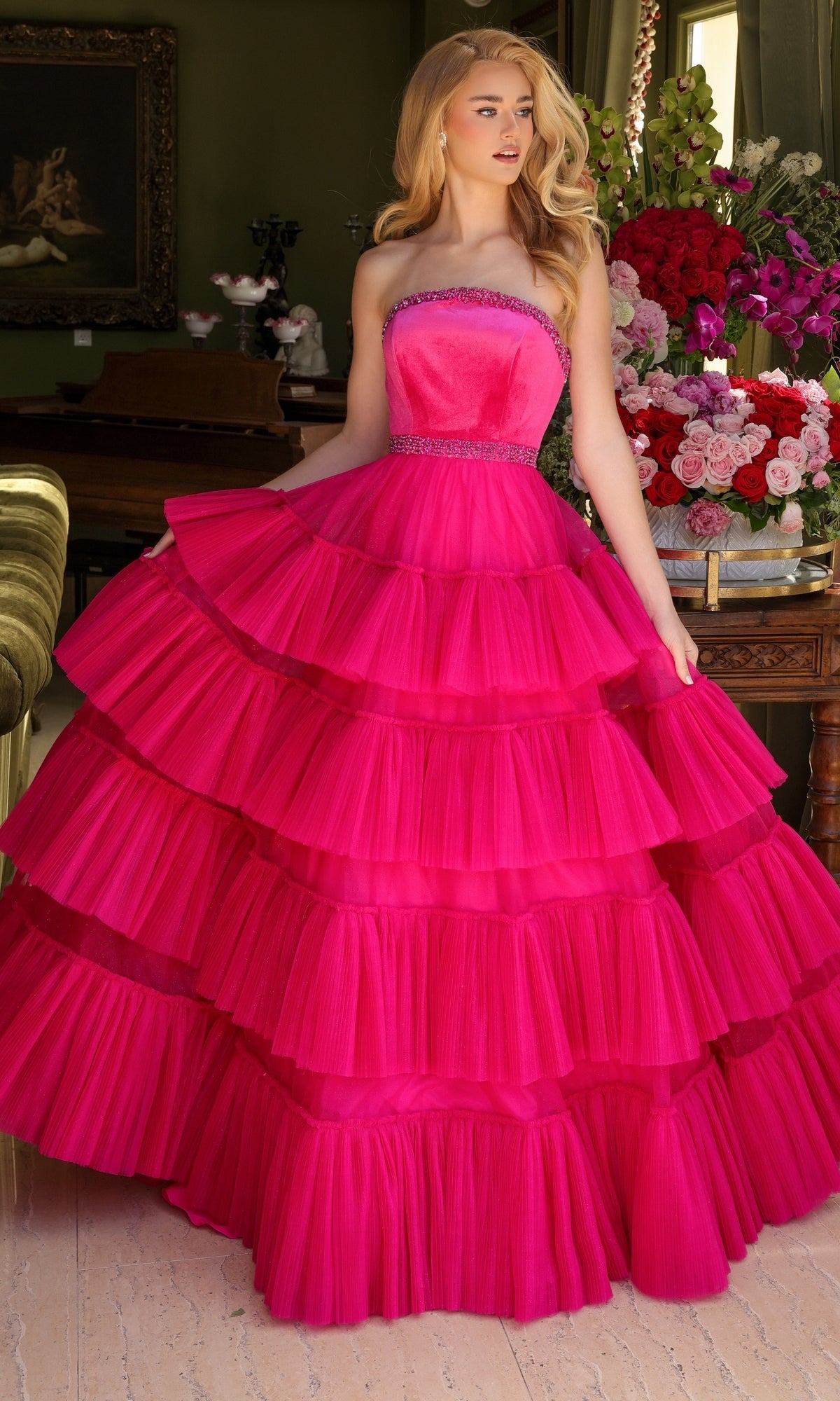 A woman stands indoors wearing the Long Prom Dress 29529 by Ava Presley, a strapless bright pink A-line dress with a multi-tiered ruffled skirt. In the background are a table with flower arrangements and a painting.
