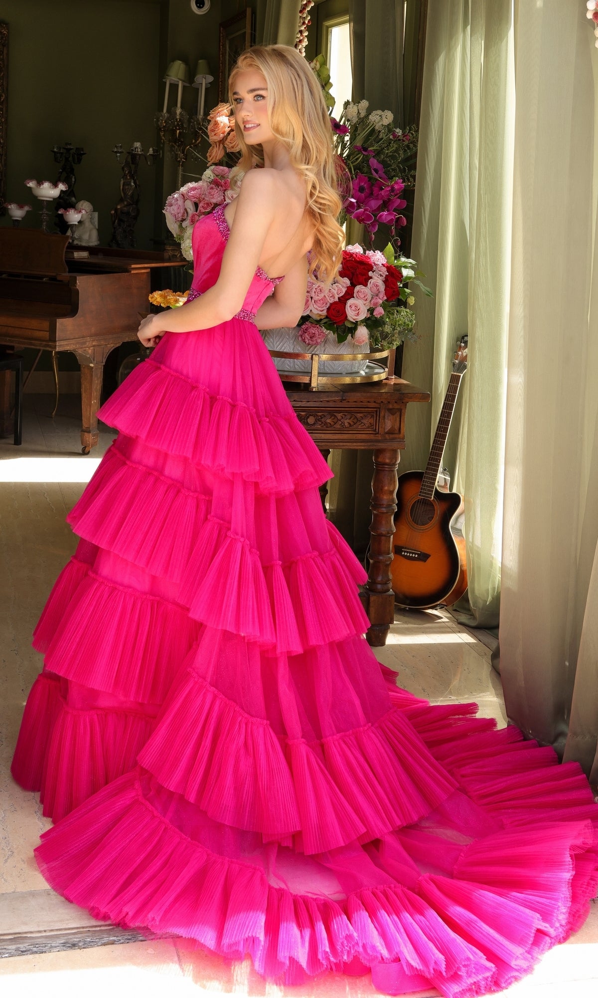 A woman with long blonde hair wears the Long Prom Dress 29529 by Ava Presley, a bright pink, layered ruffle ball gown, and stands indoors near flowers and a guitar.