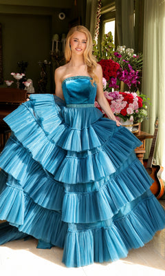 A woman stands indoors wearing the Ava Presley Long Prom Dress 29529— a strapless, layered ruffle ball gown with a beaded waist—holding one side of the skirt. Flowers and a guitar appear in the background.