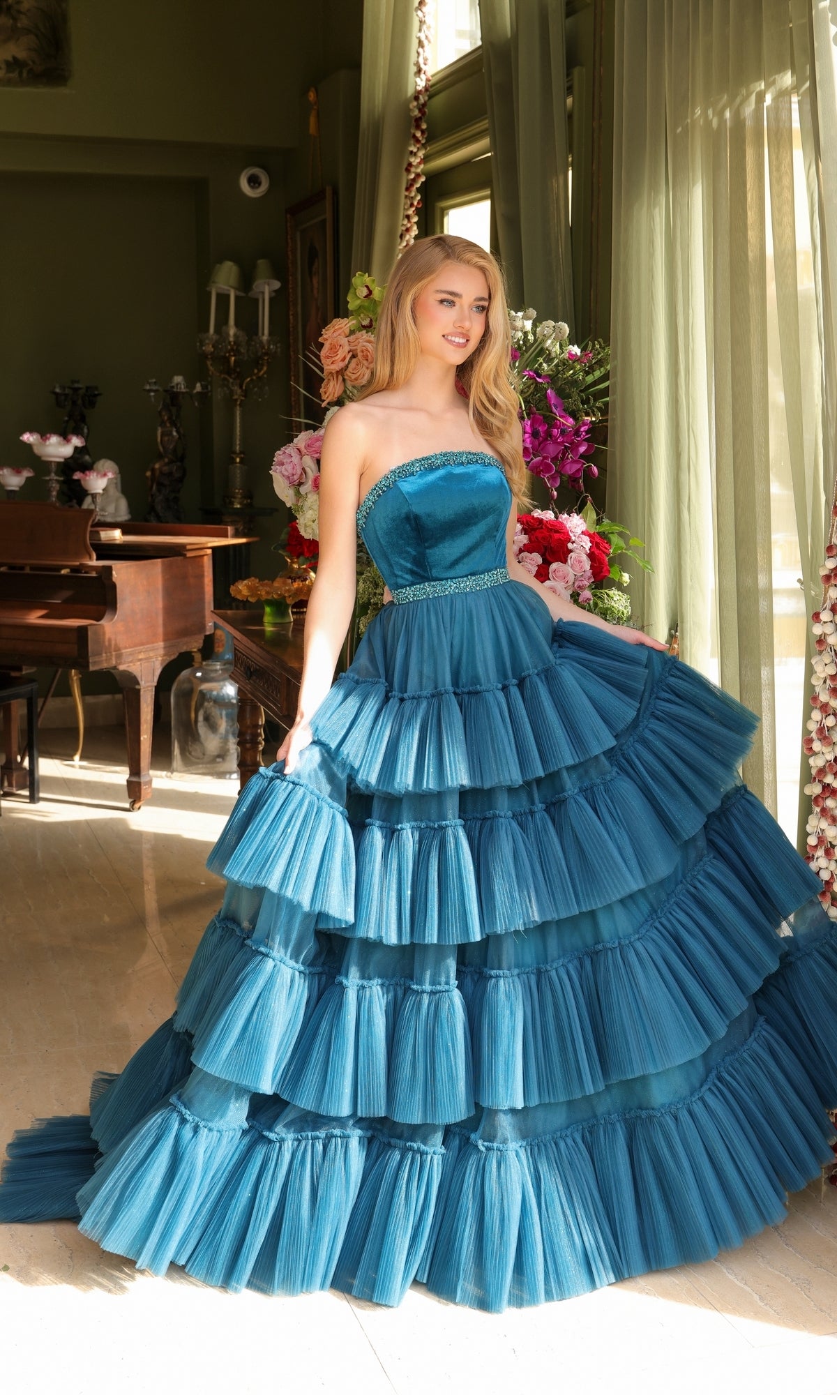 A woman stands indoors wearing the Long Prom Dress 29529 by Ava Presley, featuring a strapless bodice, beaded waist, and blue layered ruffle ball gown skirt, with flowers and a piano in the background.