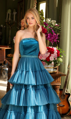 A woman wears the Long Prom Dress 29529 by Ava Presley—a strapless a-line gown with a beaded waist and tiered blue layers—standing indoors beside a flower-adorned table with a guitar resting against it.