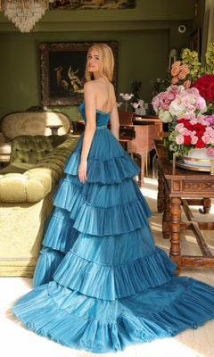 Wearing the Long Prom Dress 29529 by Ava Presley, a woman stands indoors near a green sofa and wooden table with pink and red flowers, looking over her shoulder.