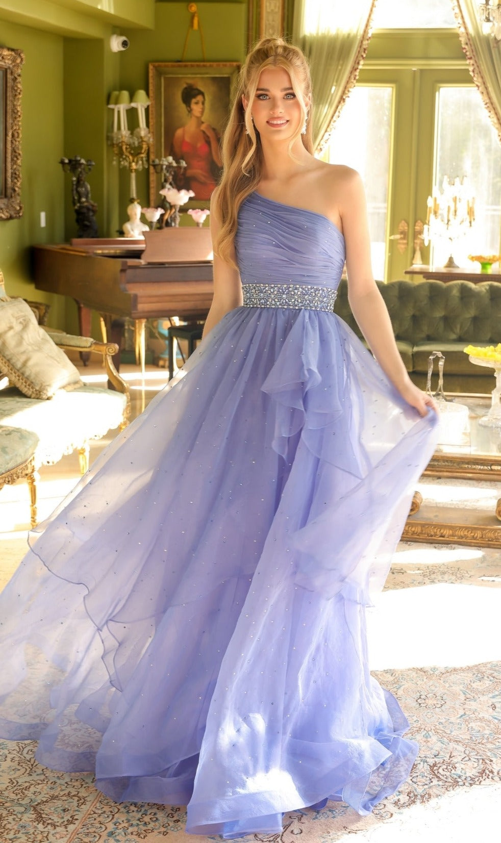 A woman stands indoors in a well-lit, elegant room wearing the Ava Presley 29534 Beaded-Waist Long Prom Dress—a floor-length, light purple gown with tulle skirt and sparkling waistband.