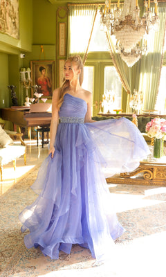 A woman in the Ava Presley 29534 beaded-waist long prom dress stands in a sunlit, elegant room with large windows, a chandelier, and classical decor.