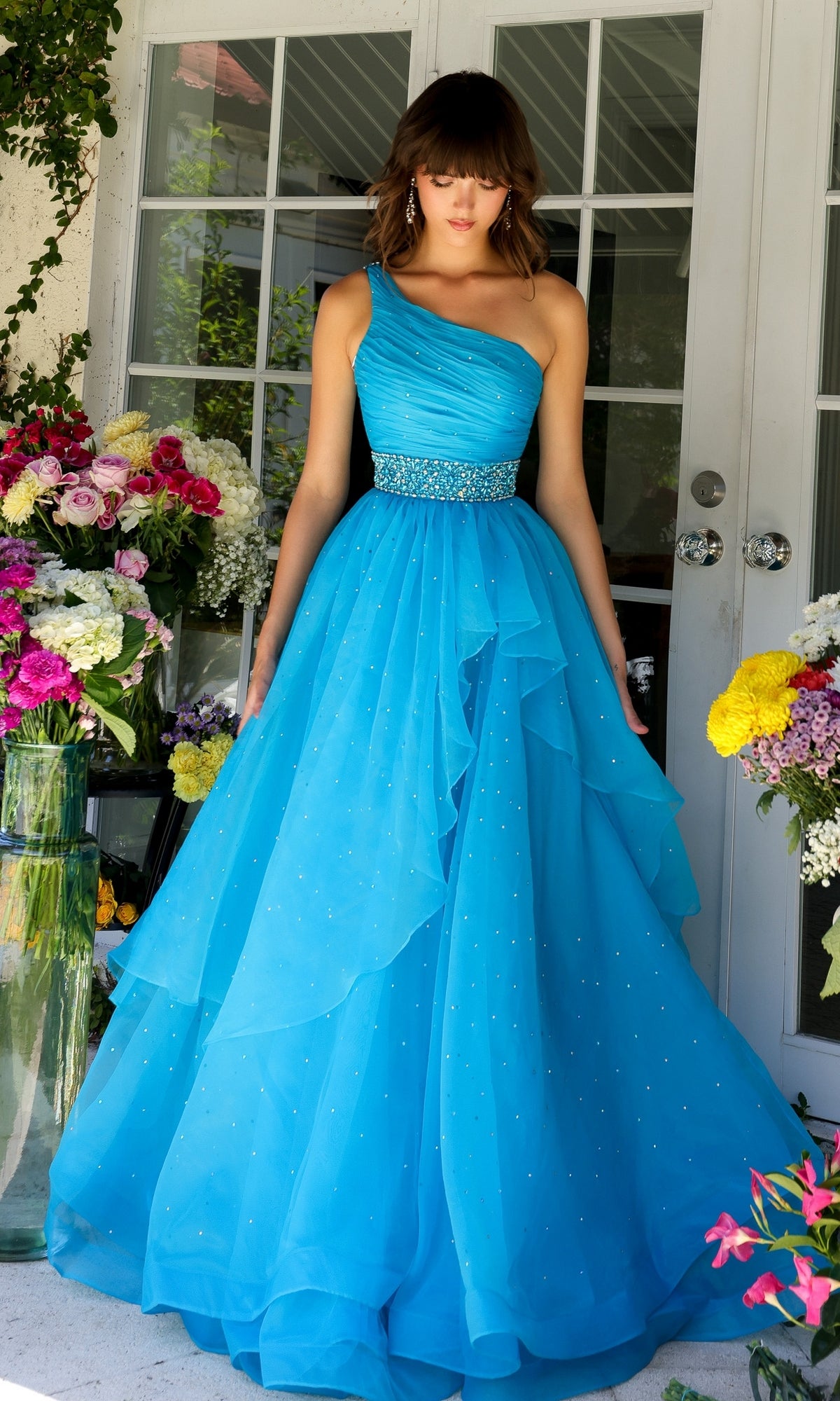 A woman in the Beaded-Waist Long Prom Dress (Ava Presley 29534) stands among colorful flower arrangements, framed by glass doors.