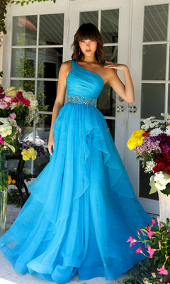 A woman wearing the Ava Presley 29534 Beaded-Waist Long Prom Dress in bright blue stands before glass doors, surrounded by colorful flowers.