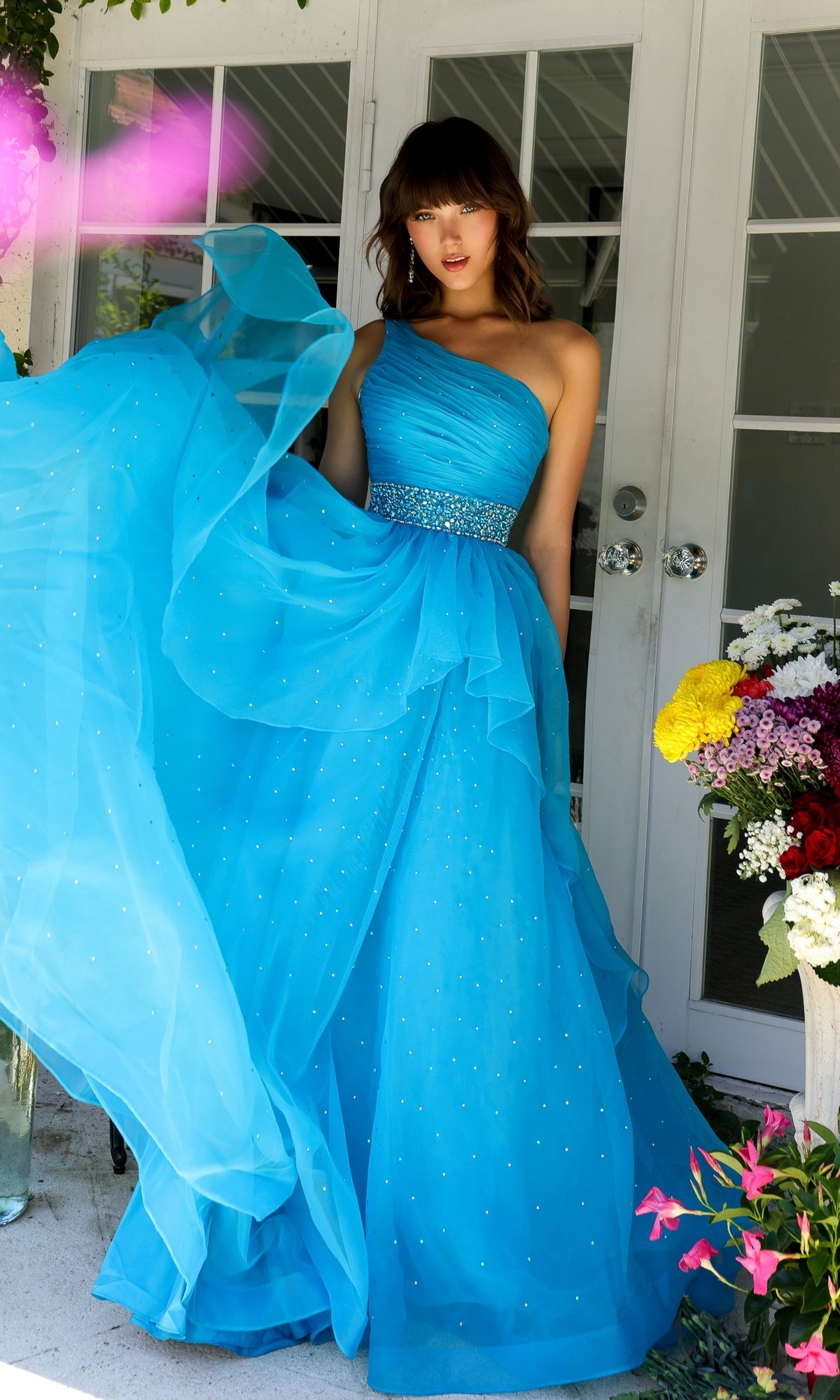 A woman in the Ava Presley 29534 Beaded-Waist Long Prom Dress stands by white doors, holding up her blue one-shoulder gown next to a vase of colorful flowers.