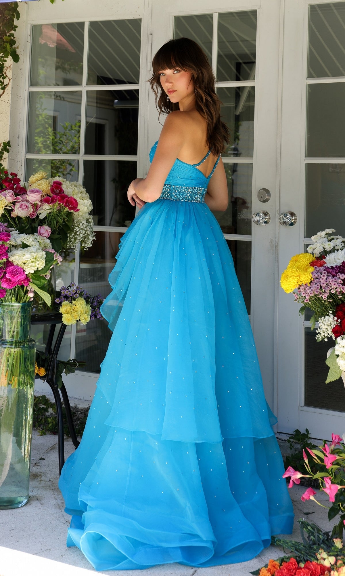 A woman in the Ava Presley 29534 Beaded-Waist Long Prom Dress stands among vibrant flowers by glass doors, glancing over her shoulder.