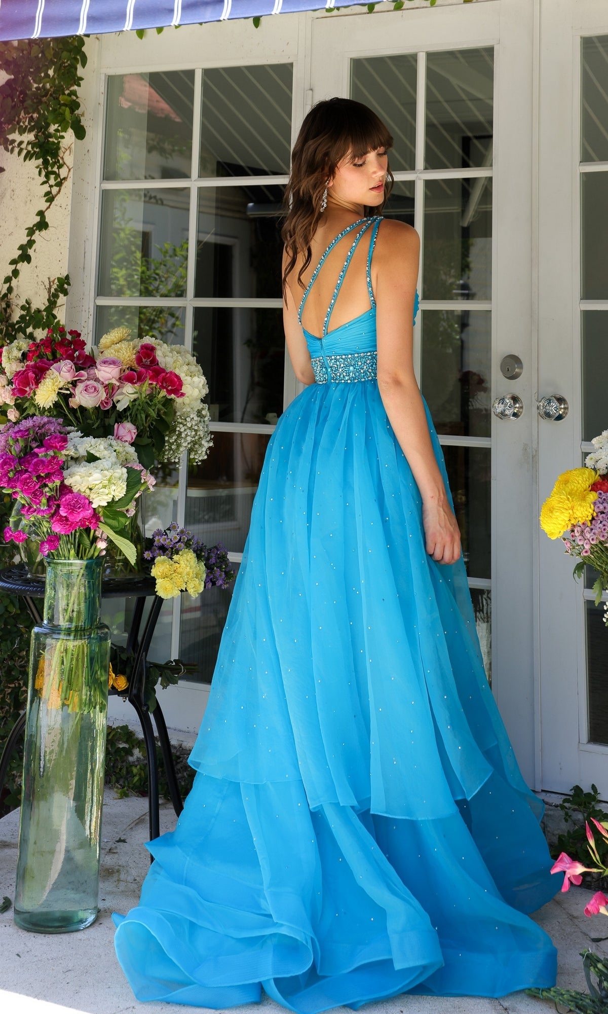 A woman in the Ava Presley 29534 Beaded-Waist Long Prom Dress stands outside by glass doors, framed by colorful floral arrangements.