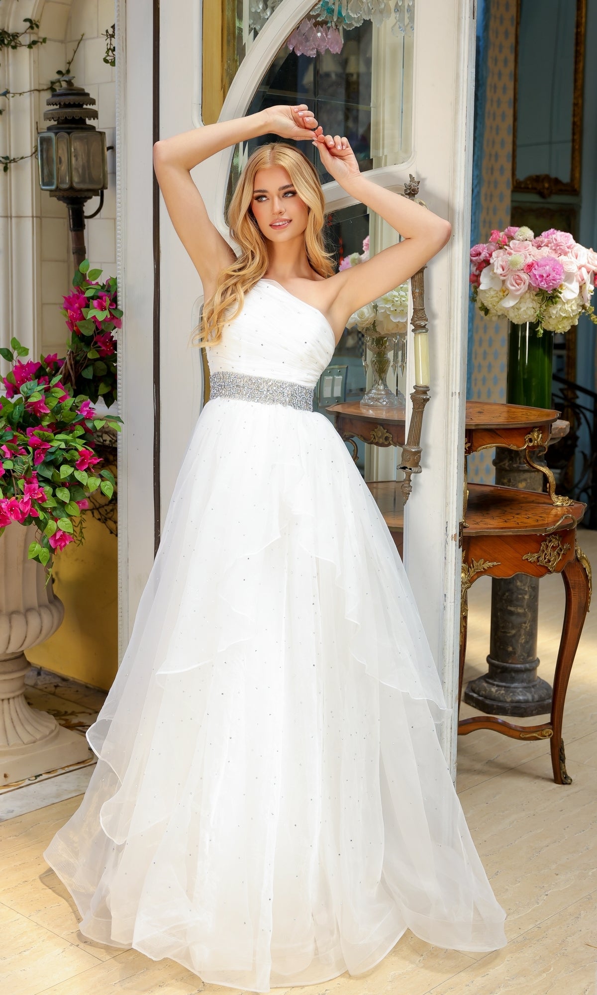 A woman wearing the Beaded-Waist Long Prom Dress by Ava Presley 29534 stands in a doorway, flowers and antique furniture around her.