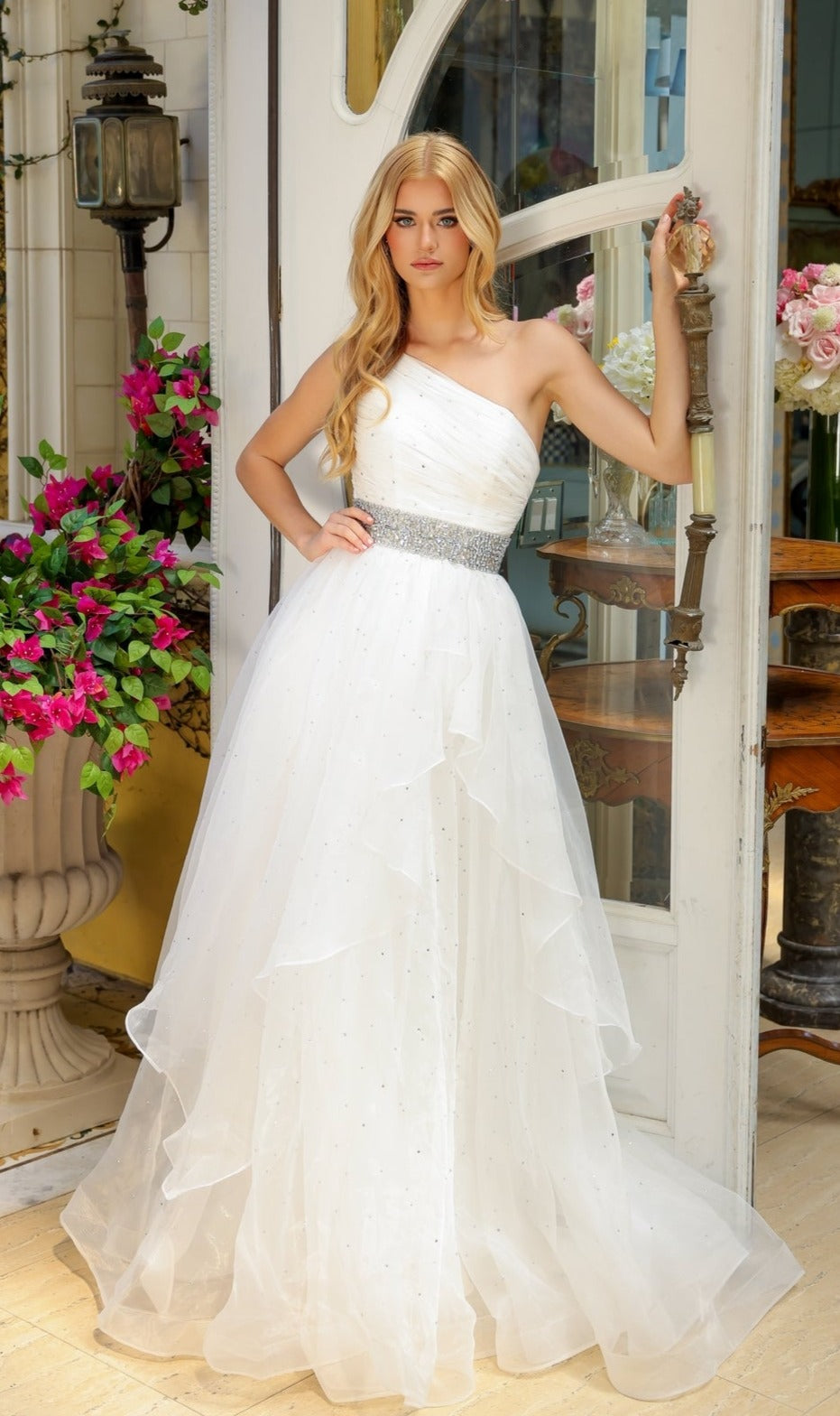A woman in an Ava Presley 29534 Beaded-Waist Long Prom Dress stands by an open door, surrounded by potted flowers and indoor plants.