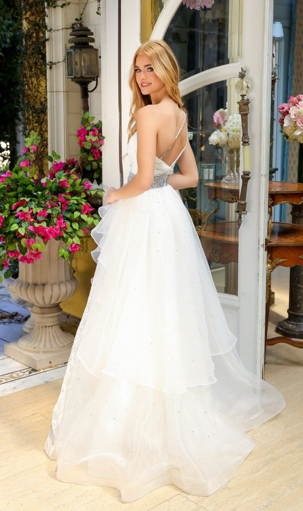 A woman in the Beaded-Waist Long Prom Dress by Ava Presley 29534, featuring layered white fabric and thin straps, stands near a doorway indoors with pink flowers and elegant decor in the background, looking back over her shoulder.