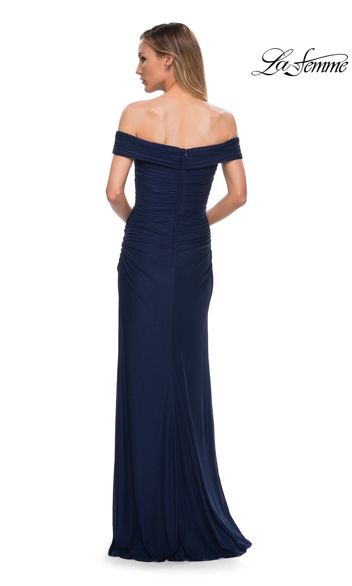 A woman models the Long Formal Dress: La Femme 29541 from the back, showcasing its floor-length navy blue design with an off-the-shoulder neckline and ruched bodice.