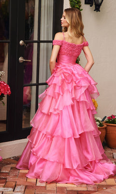 A woman stands on a brick patio facing away, wearing the Ava Presley 29544 Ruffled Prom Ball Gown with off-the-shoulder straps and a princess a-line skirt, in front of glass doors and potted plants.