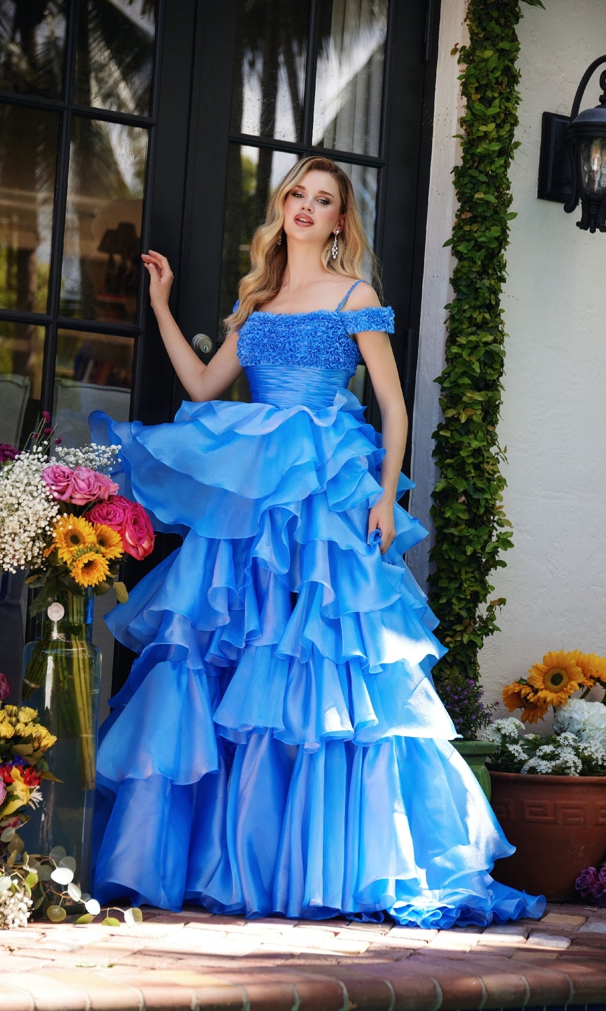 A woman wearing the Ava Presley 29544 Ruffled Prom Ball Gown with off-the-shoulder straps stands by a glass door, surrounded by potted flowers and lush greenery.