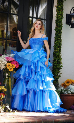 A woman wearing the Ava Presley 29544 Ruffled Prom Ball Gown with off-the-shoulder straps stands by a glass door, surrounded by potted flowers and lush greenery.