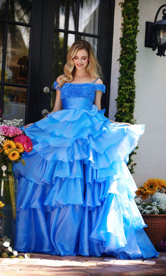 A woman wearing the Ava Presley 29544 ruffled blue prom ball gown with off-the-shoulder straps stands outside near glass doors, surrounded by sunflowers and colorful flowers.