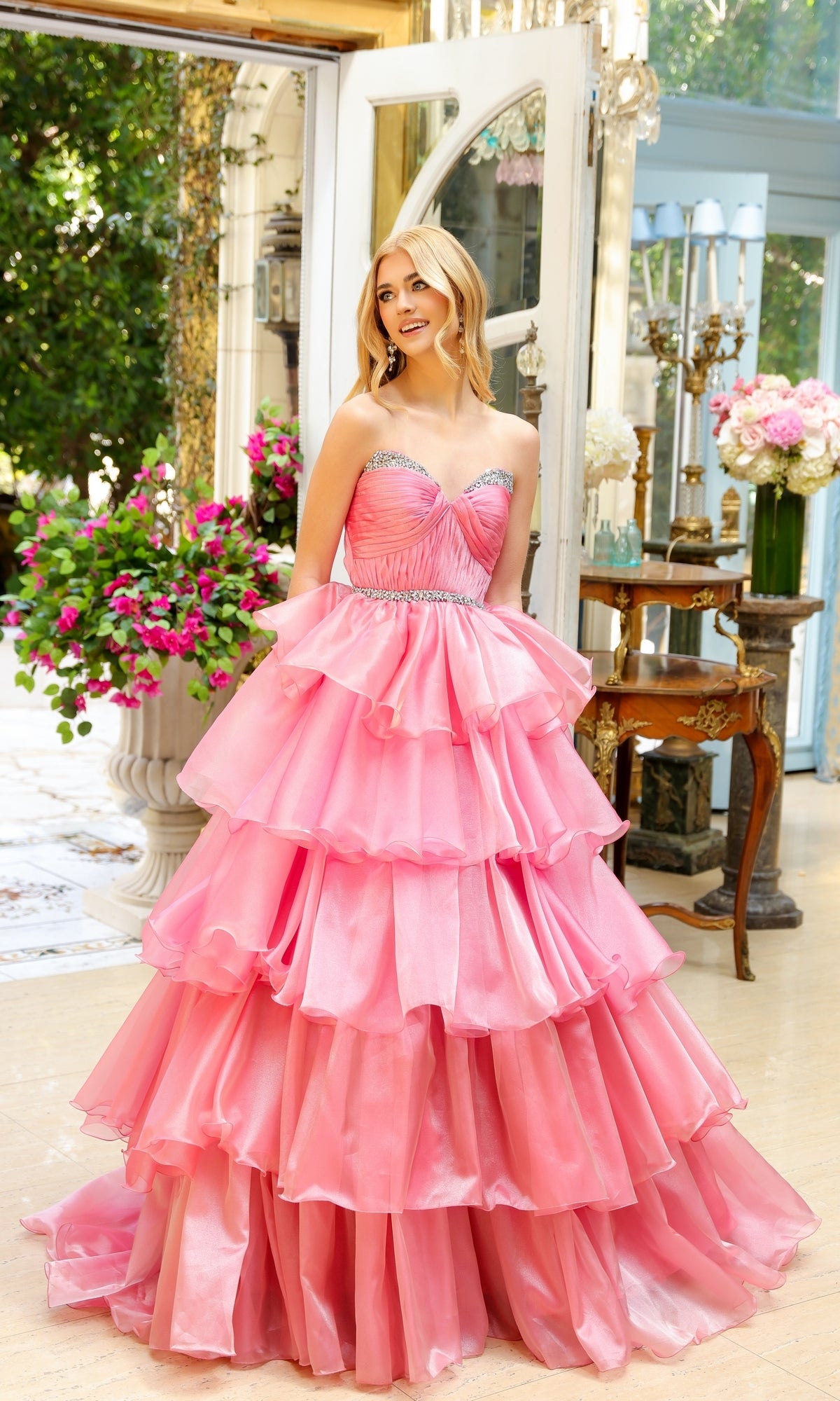 A woman smiles indoors surrounded by ornate furniture and vibrant flowers, wearing the Long Prom Dress 29546 by Ava Presley—a pink strapless ball gown with a sweetheart neckline and layered ruffles.