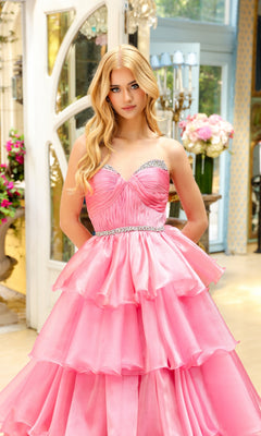 A woman with long blonde hair wears the Long Prom Dress 29546 by Ava Presley, a strapless pink ruffle ball gown with a jeweled waistline, standing indoors near floral arrangements and large windows.