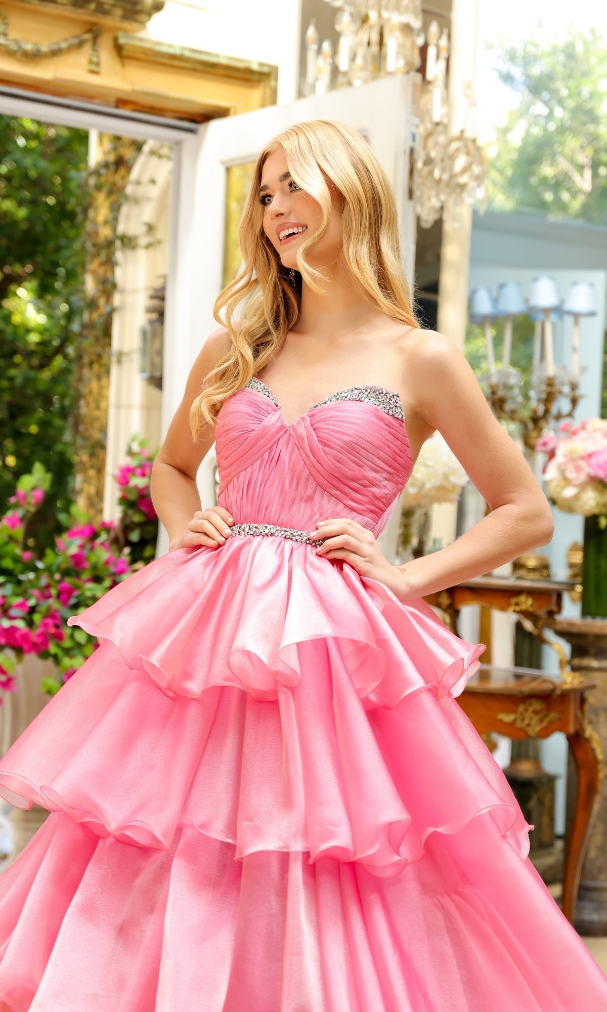 A woman stands indoors in the Long Prom Dress 29546 by Ava Presley, featuring a strapless sweetheart neckline, layered pink ball gown design with rhinestone accents, surrounded by flowers and elegant decor.