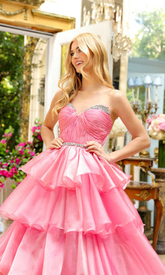 A woman stands indoors in the Long Prom Dress 29546 by Ava Presley, featuring a strapless sweetheart neckline, layered pink ball gown design with rhinestone accents, surrounded by flowers and elegant decor.