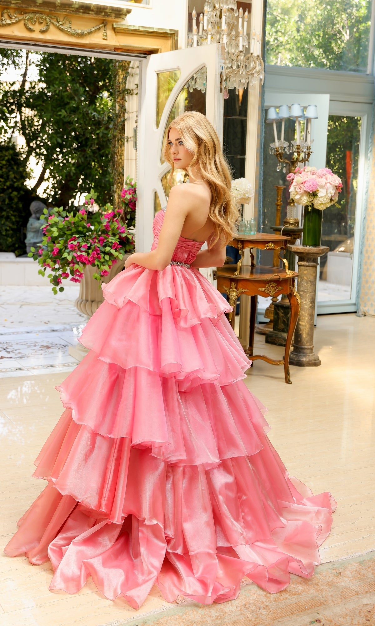 A woman wearing the Long Prom Dress 29546 by Ava Presley, featuring a strapless sweetheart neckline and layered ruffle ball gown, stands indoors by open doors with flowers and elegant furniture around her.
